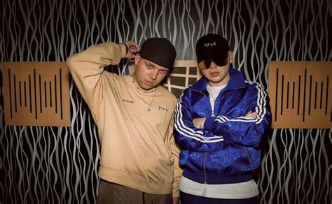 dos artistas, Bizarrap y Arcángel, en un estudio de grabación, con Bizarrap detrás de la consola y Arcángel de pie, ambos con expresiones de concentración y energía.